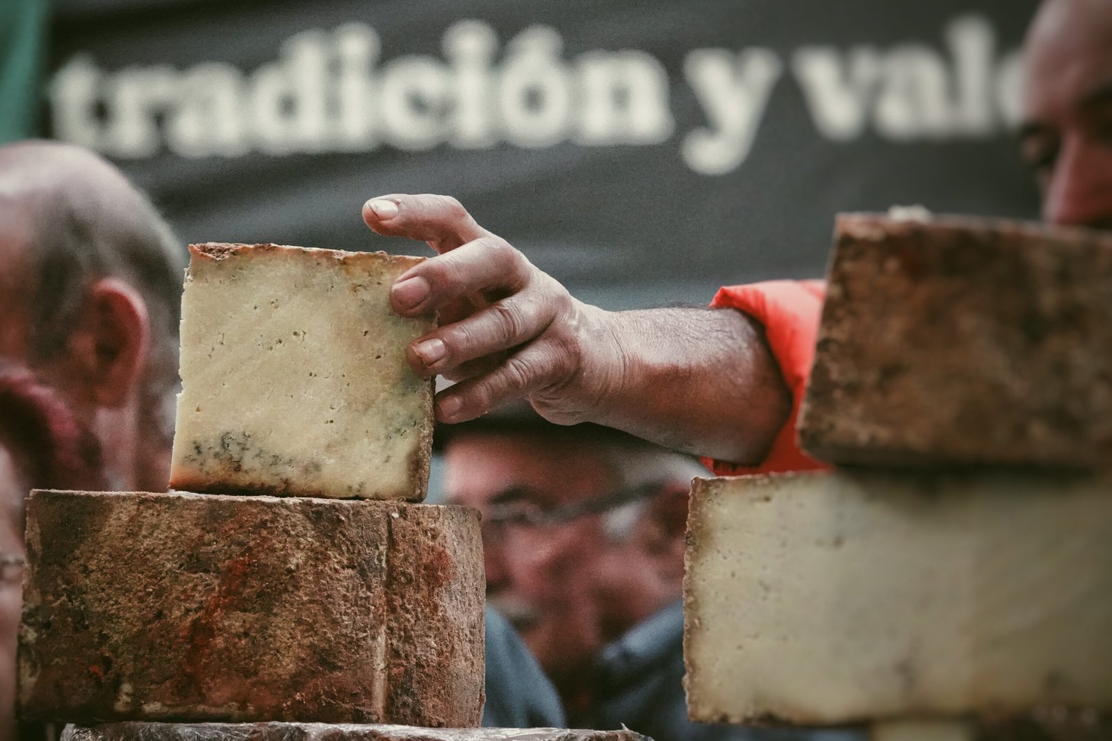 Mano de un vendedor sosteniendo un bloque de queso en un puesto de feria, con una rueda de queso y más piezas al lado