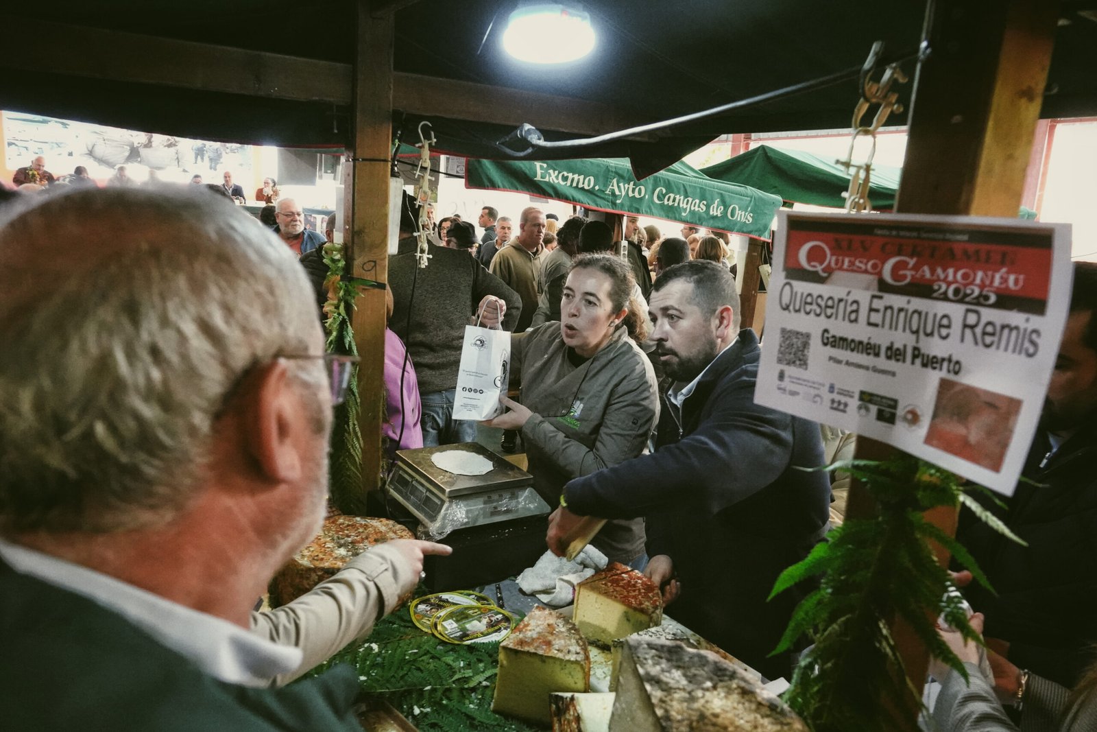 Puesto de la Quesería Enrique Remis en el XLV Certamen del Queso Gamonéu, con dos dependientes tras el mostrador y quesos expuestos sobre hojas de helecho