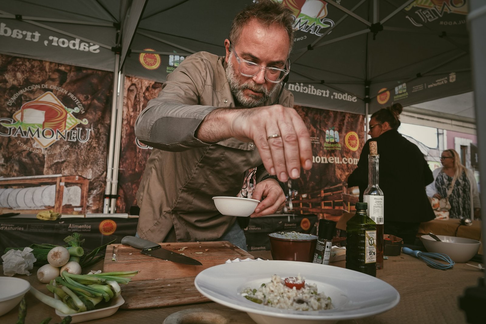 Cocinero aliñando un plato en una demostración culinaria bajo una carpa, con lonas del Gamonéu DOP al fondo