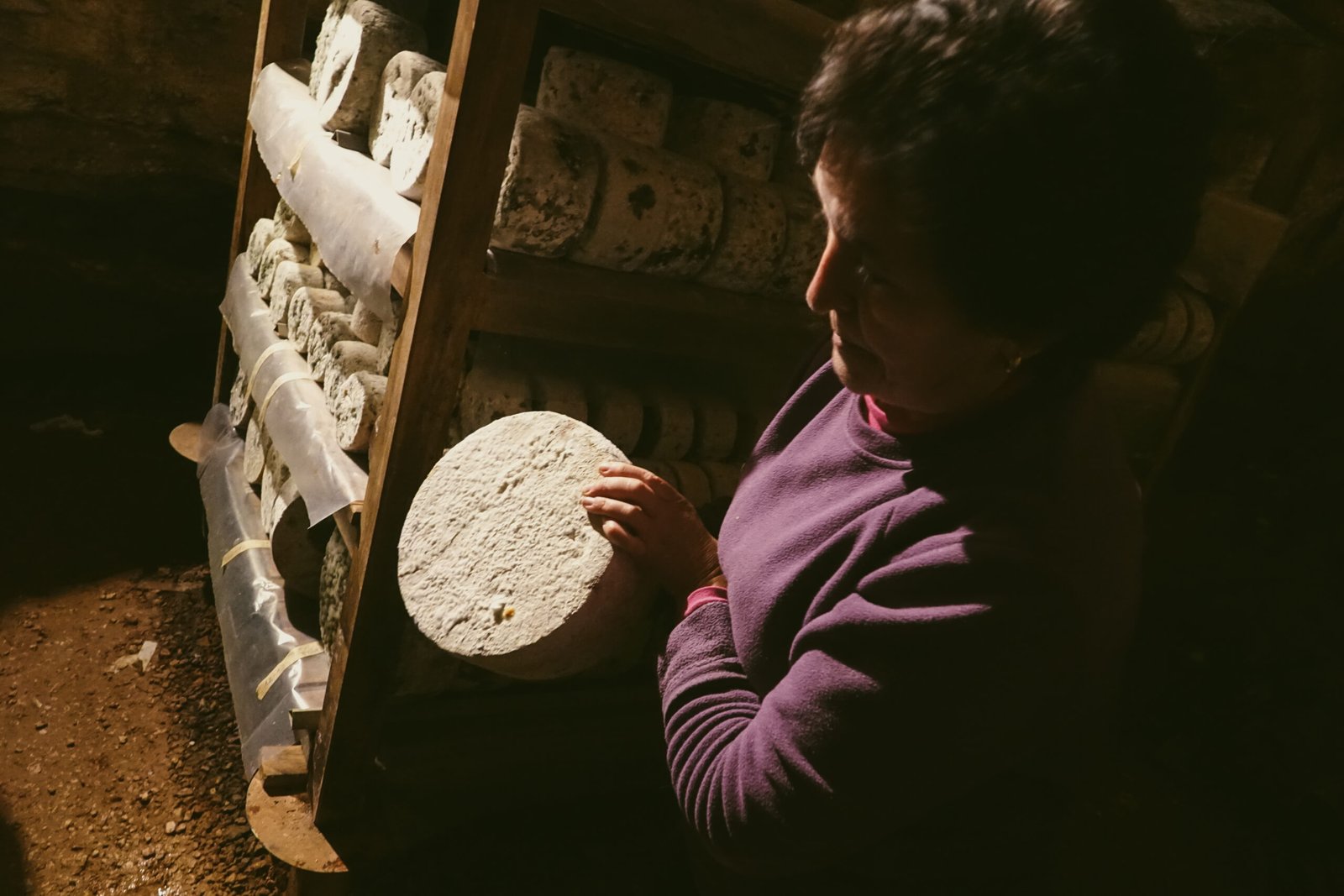 Mujer de perfil sosteniendo un queso curado junto a una estantería de madera llena de quesos en la cueva